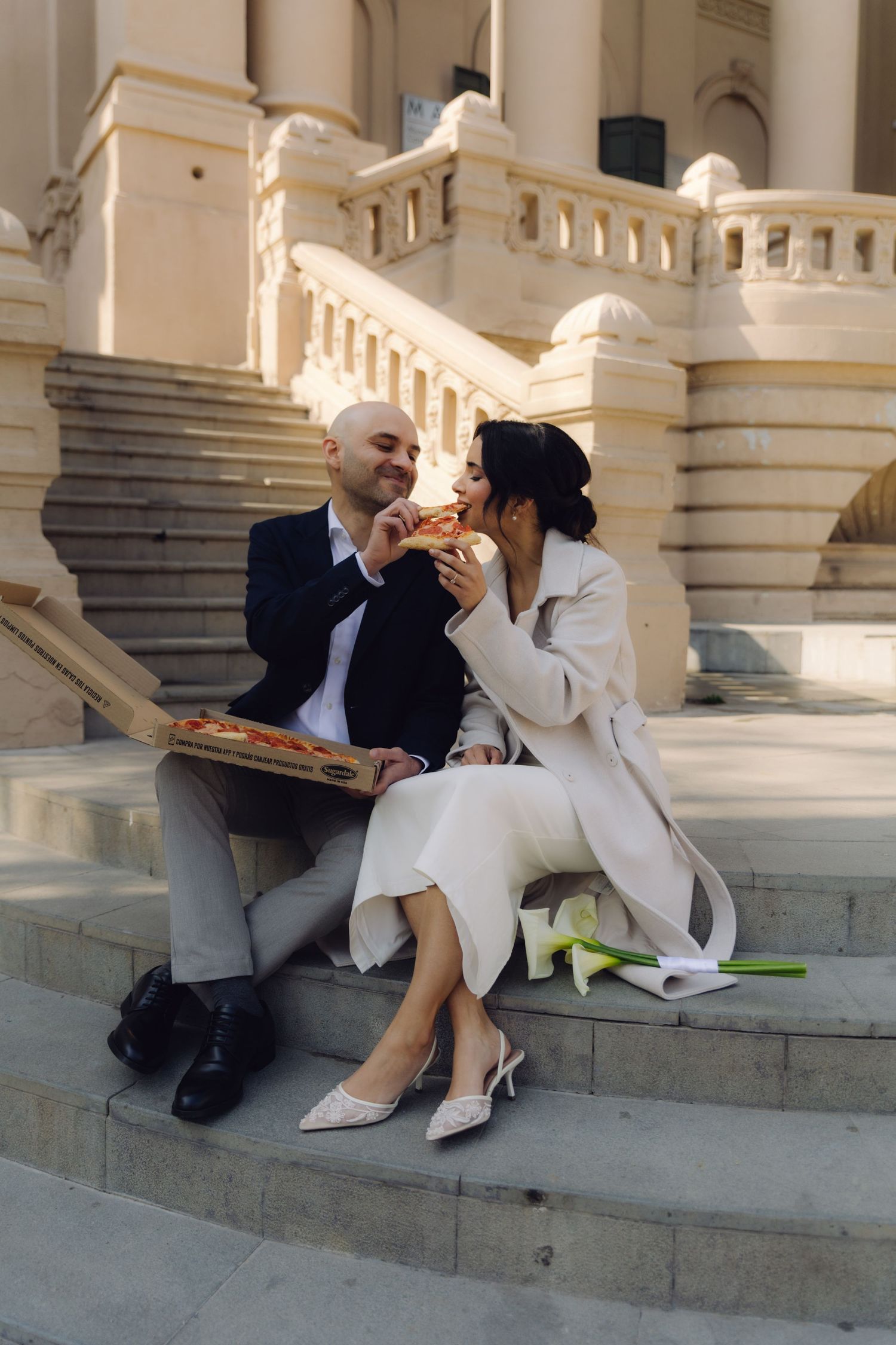 Bride and groom in formal attire sit on stone steps sharing pizza outside a classical building.