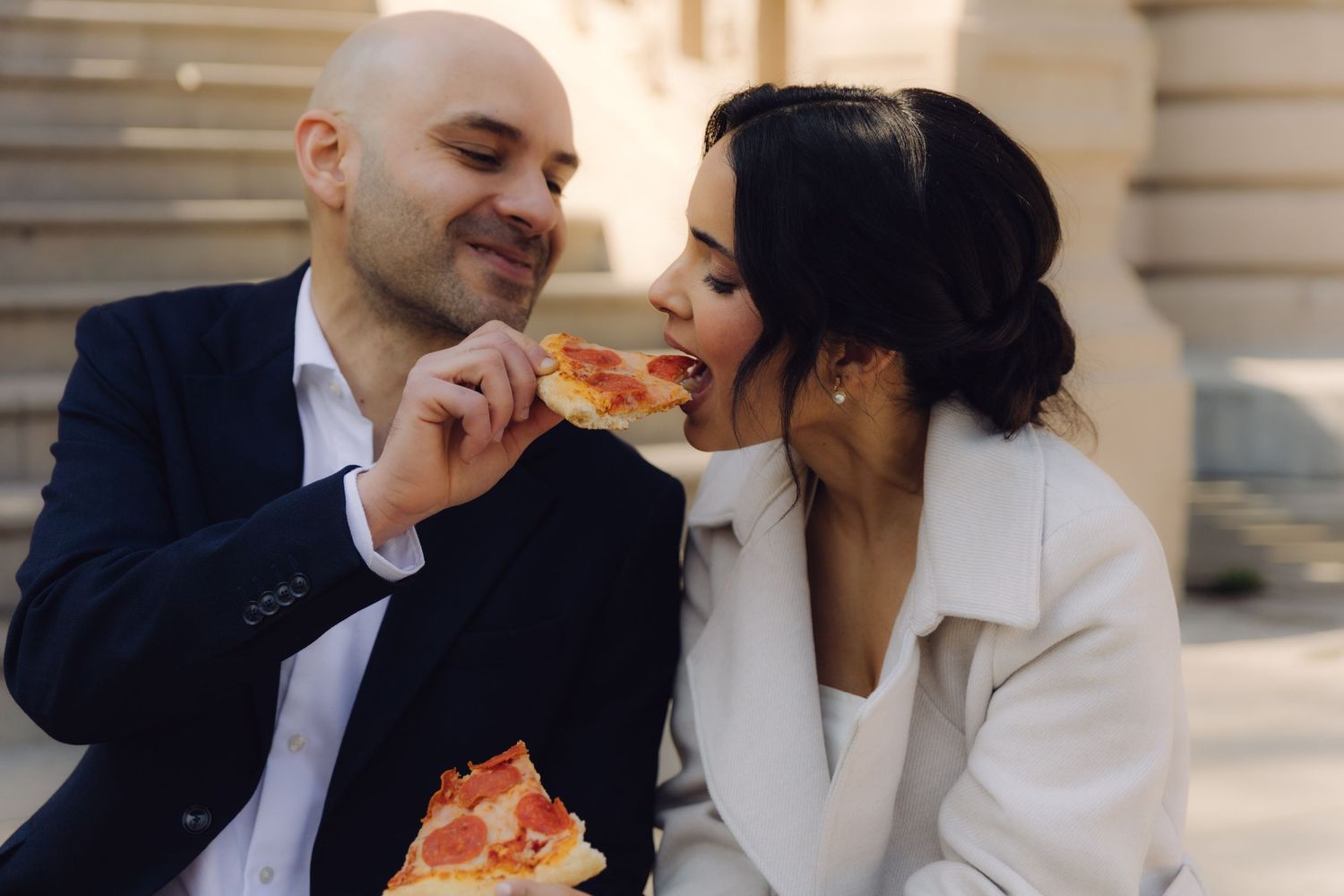 Smiling groom feeds pizza to his bride during a casual outdoor wedding celebration on city steps.
