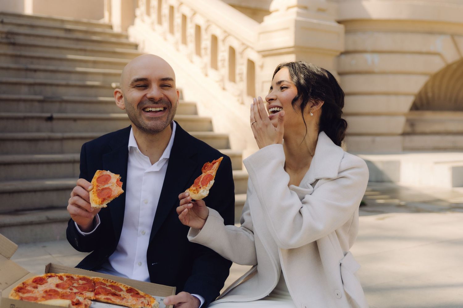 Laughing newlyweds enjoy pizza slices outdoors on the steps of a grand neoclassical building.