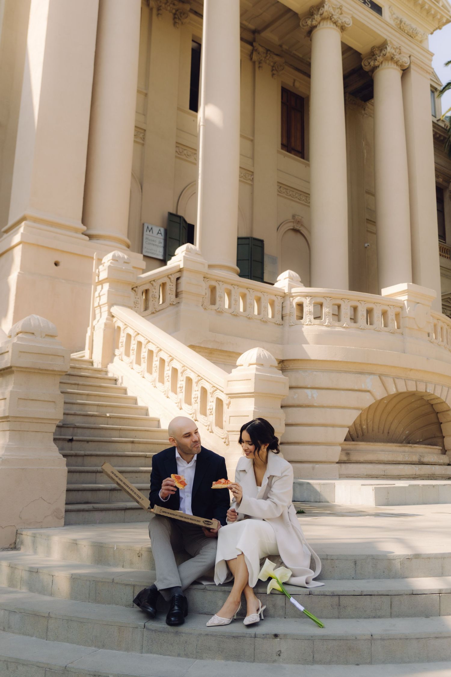 Bride and groom sit on grand stone steps of a neoclassical building sharing a pizza box.