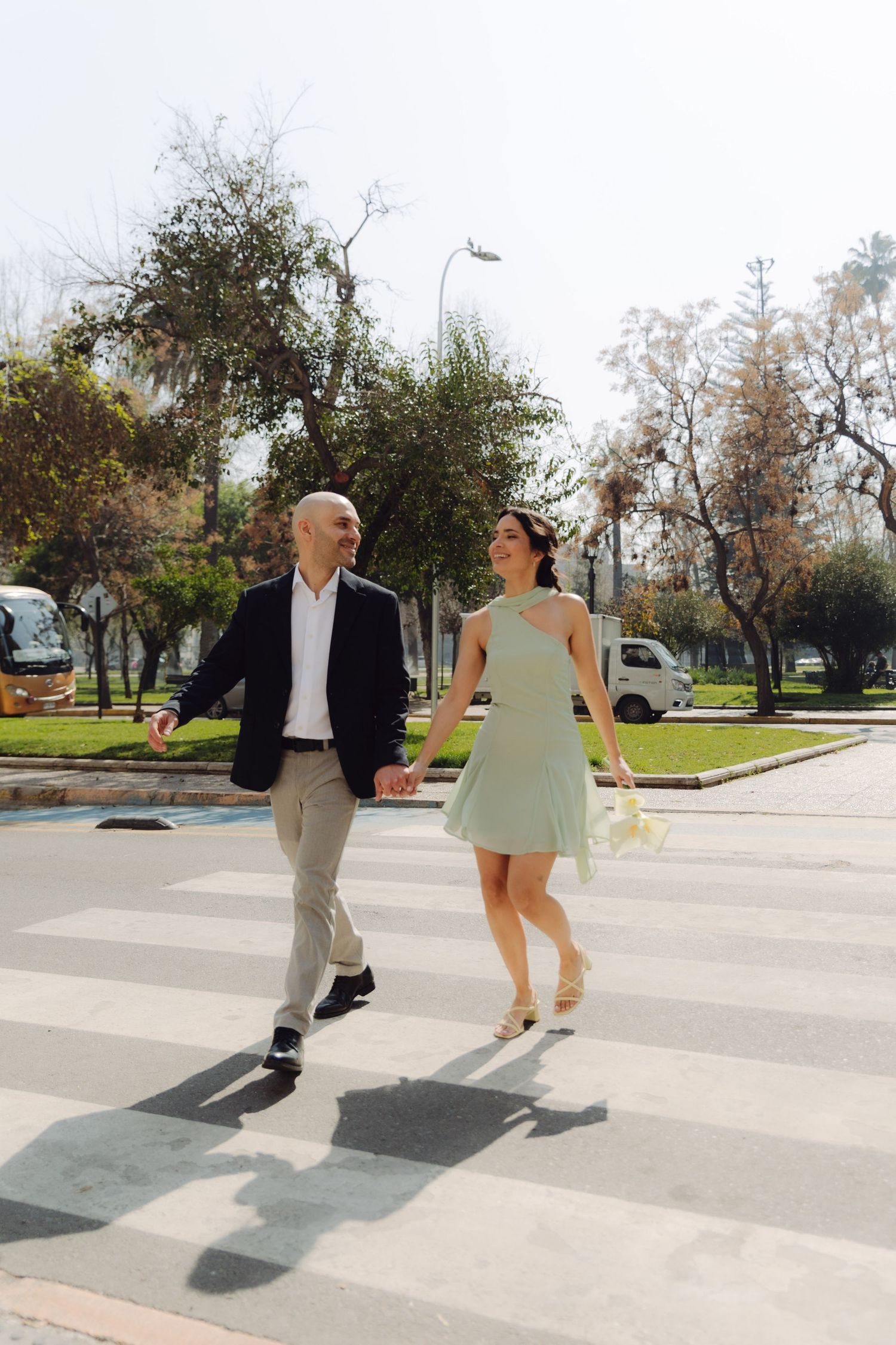 Happy couple holding hands crossing a zebra crossing in a sunny urban park after their wedding.