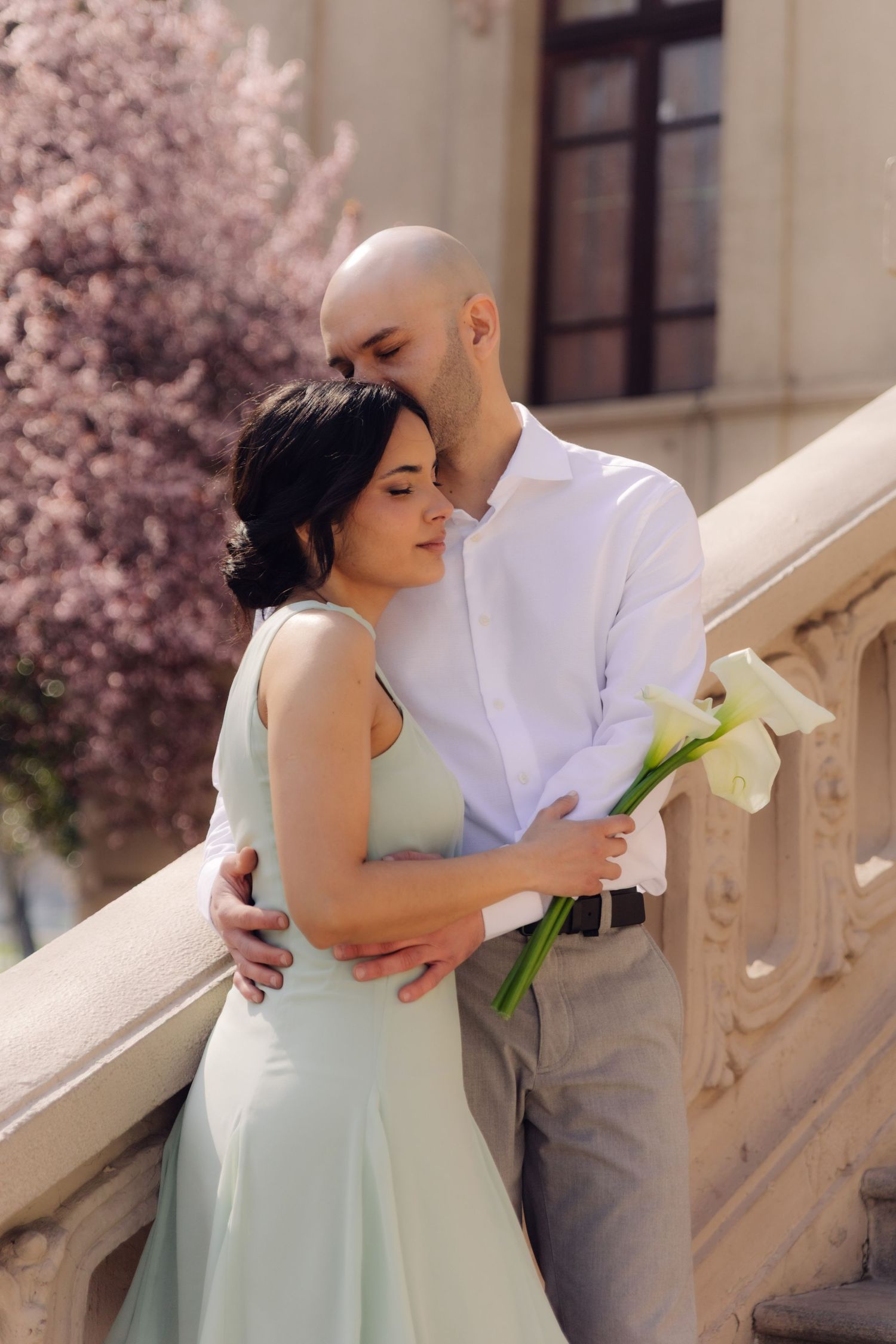 Romantic couple embracing on elegant stone stairs with cherry blossoms, woman in mint dress holding white calla lilies.