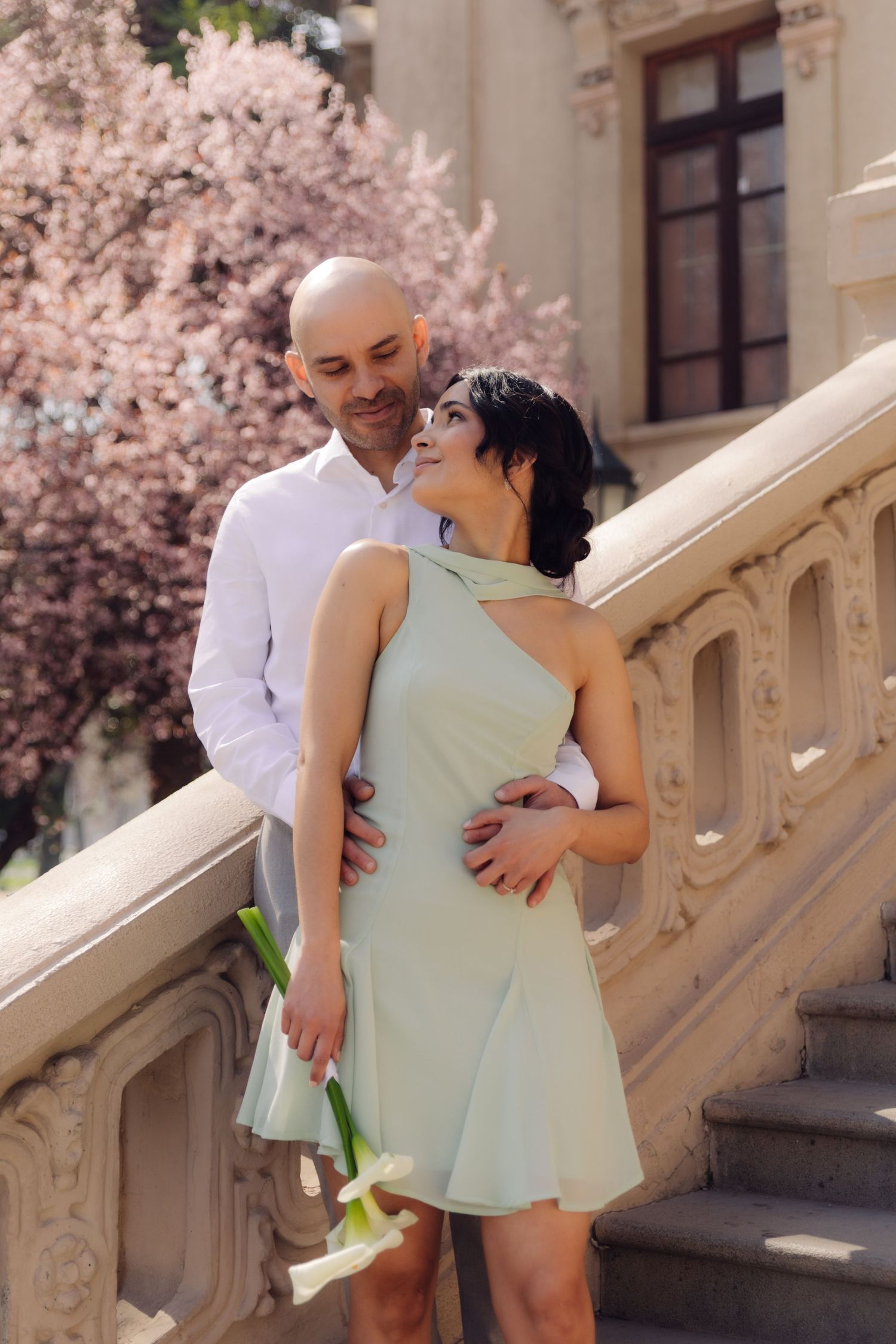 Couple sharing tender moment on historic stone steps, woman in mint dress holding calla lilies with cherry blossoms behind.