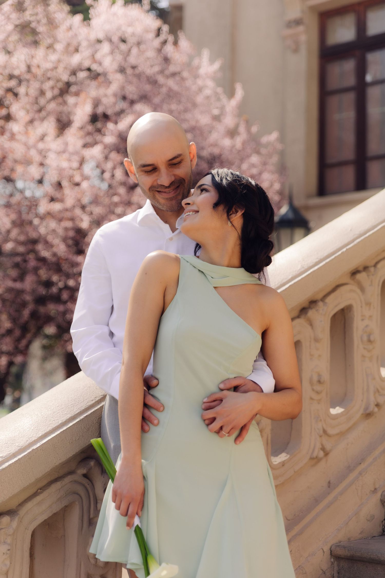 Happy couple embracing on stone balustrade, woman in mint dress laughing with pink cherry blossom trees in background.