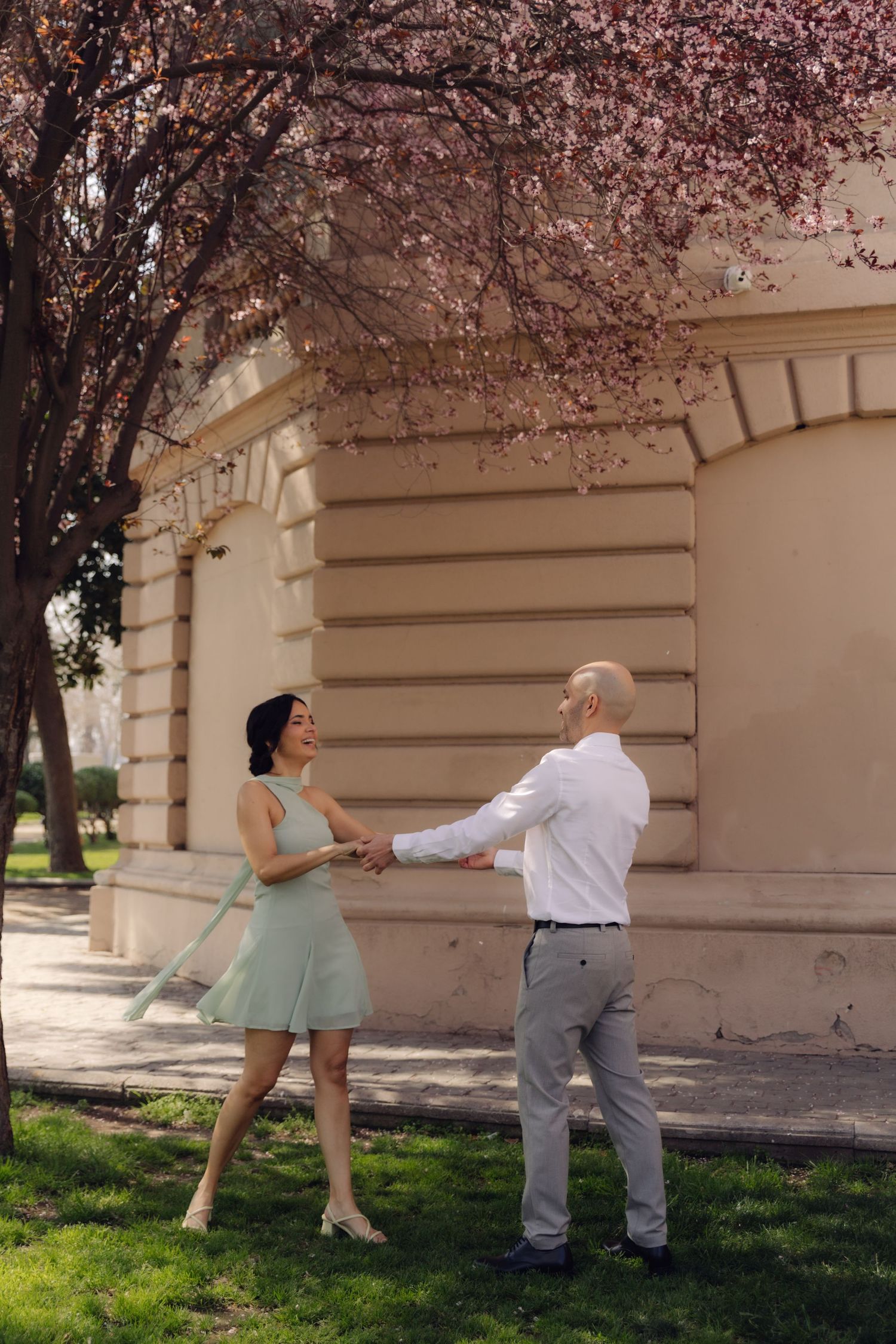 Joyful couple holding hands and dancing on green grass near blooming pink cherry tree by classic building exterior.