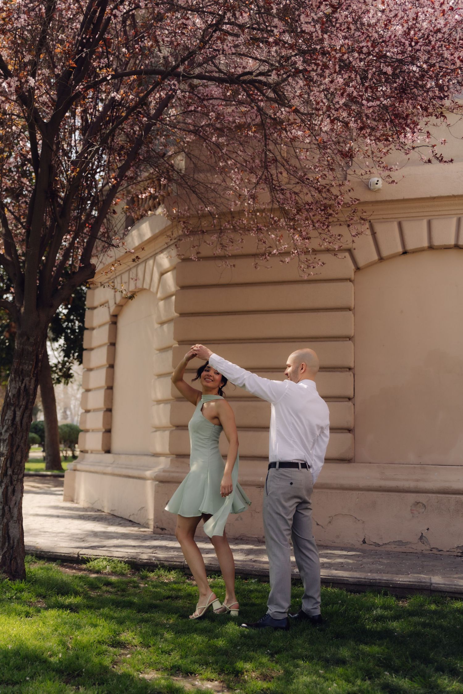 Bald man spinning dark-haired woman in mint dress under pink flowering cherry blossom tree near elegant building.