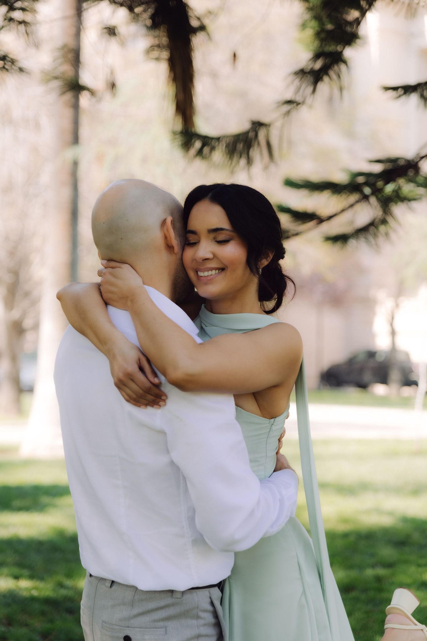 Happy woman in mint halter dress smiling while embracing bald man in white shirt among tall park trees.