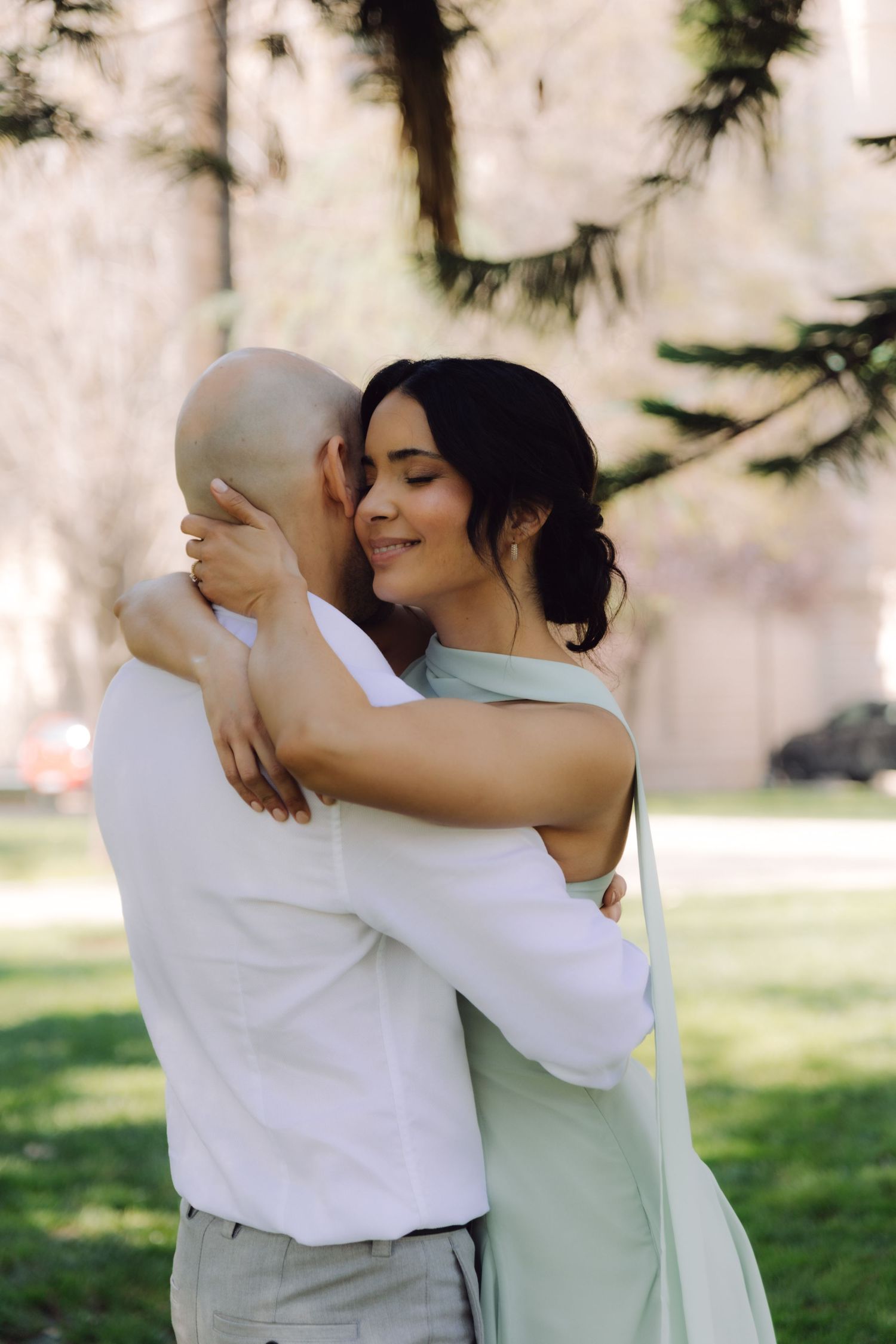 Smiling dark-haired woman in mint dress embracing bald man in white shirt in sunny green park setting.