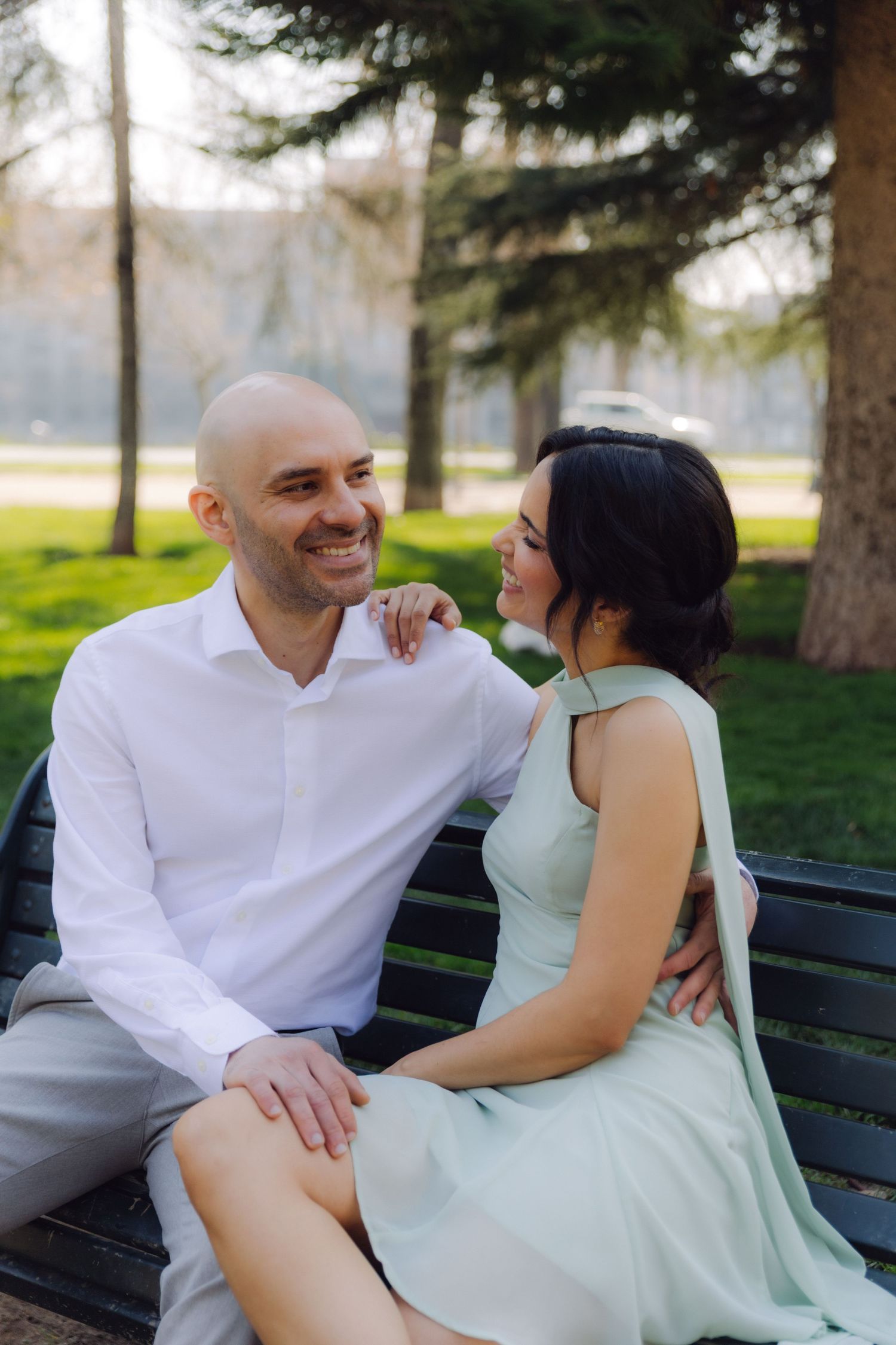 Smiling bald man and dark-haired woman in mint dress sitting together on park bench surrounded by tall trees.