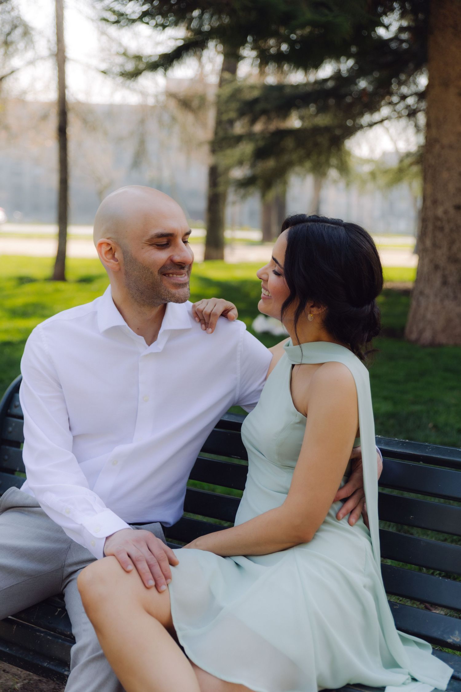 A couple sits intimately on a park bench, smiling at each other, man in white shirt and woman in mint green dress.
