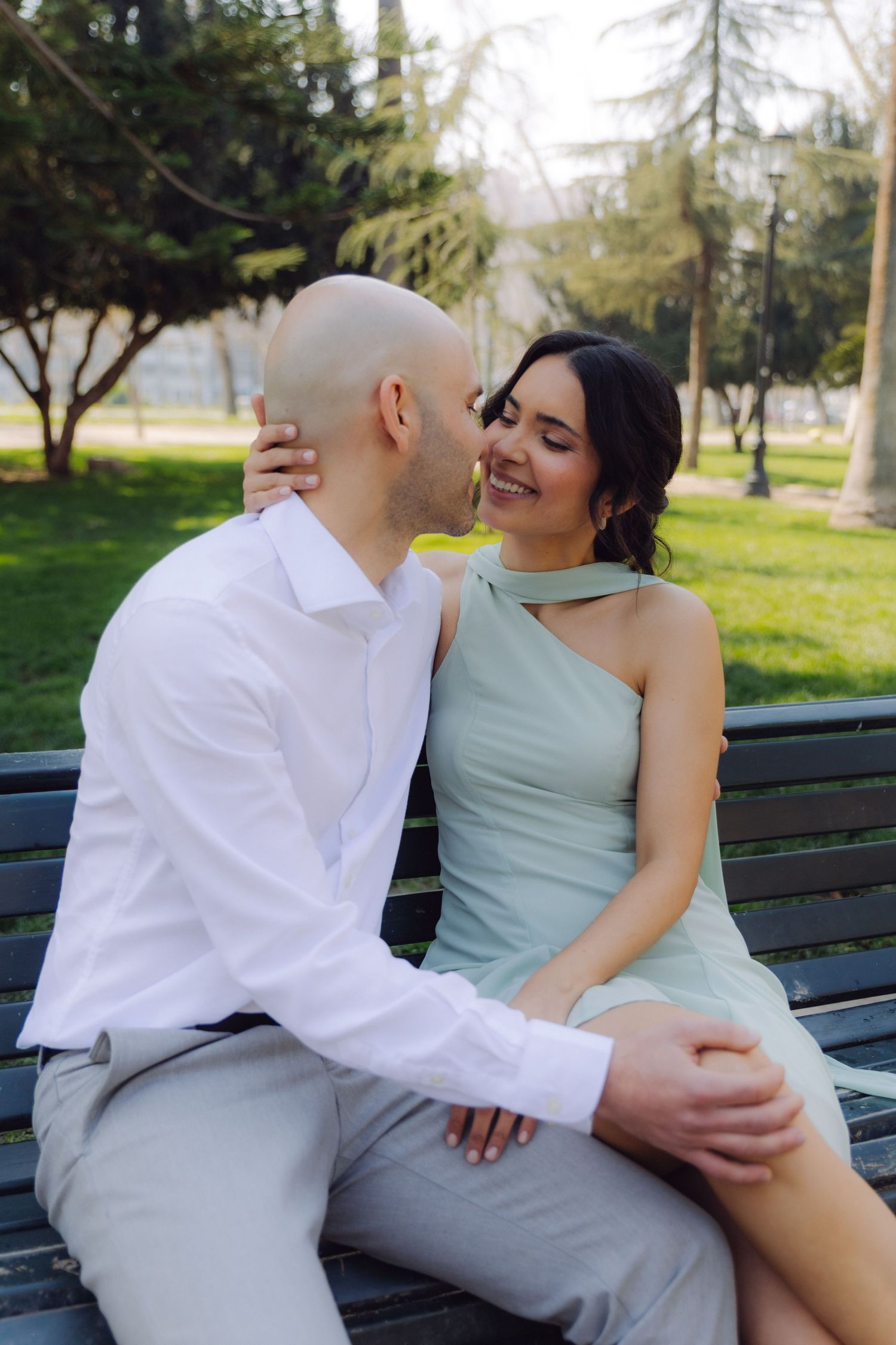 A happy couple shares a romantic moment on a park bench, woman in mint dress laughing as man leans in close.