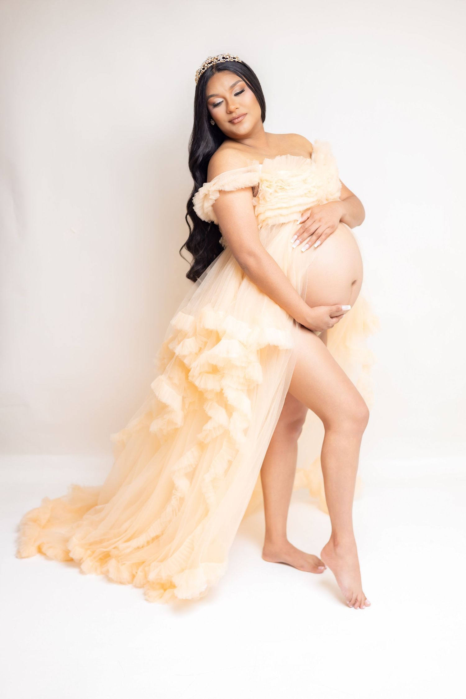Pregnant woman in elegant gold ruffled maternity gown with tiara, cradling baby bump in white studio.