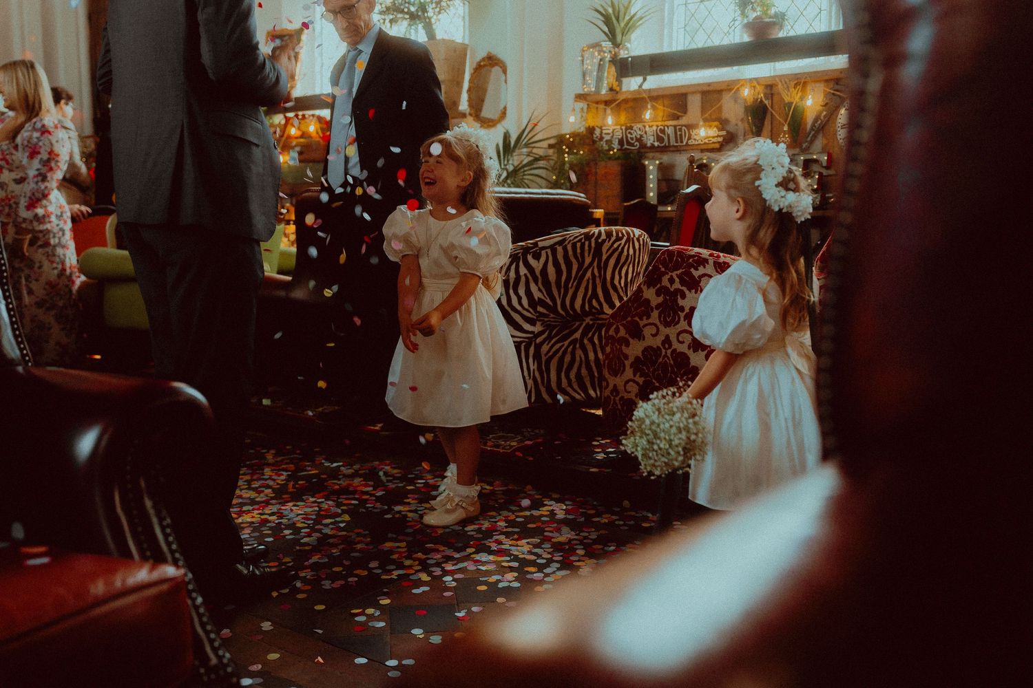 Two young flower girls in white dresses playing with colorful confetti during a wedding ceremony indoors.