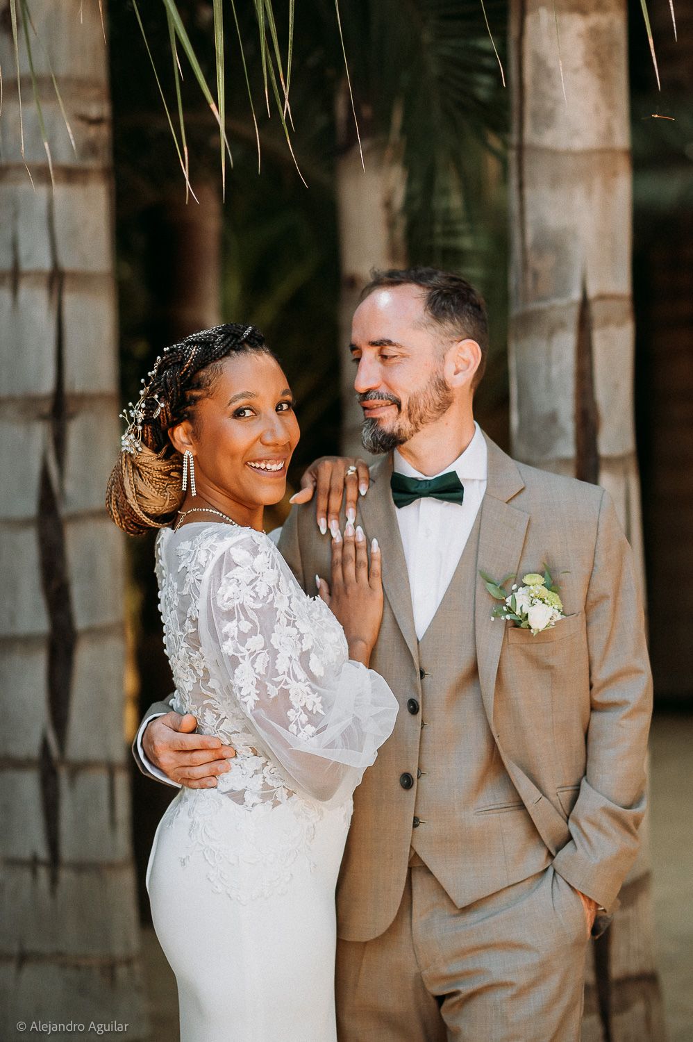 A smiling bride in lace gown and groom in tan suit with bow tie pose together at an outdoor wedding.