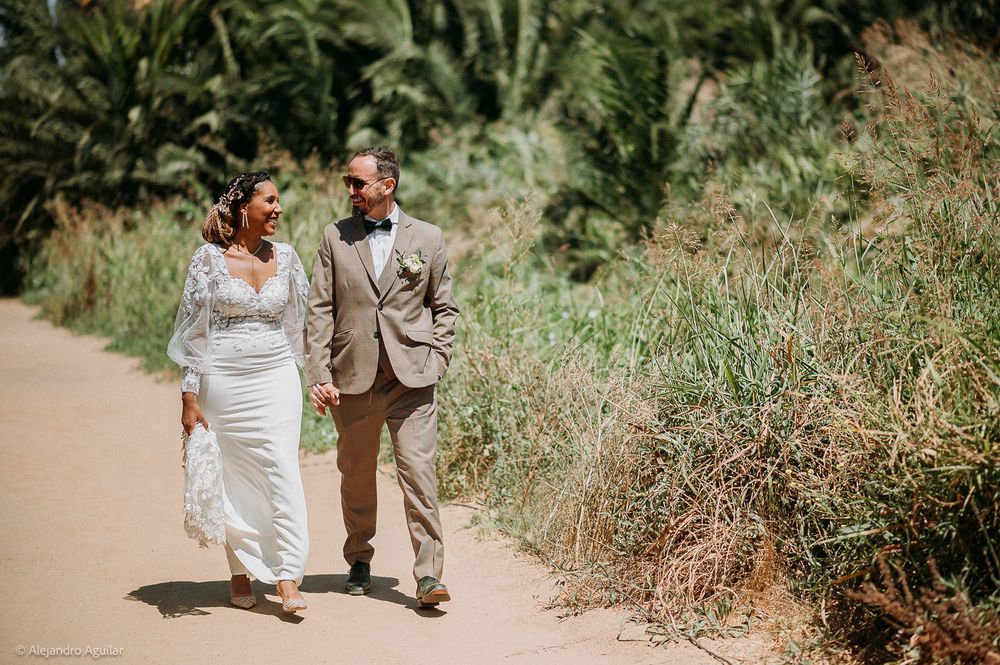 Bride and groom walking hand in hand along a tropical dirt path surrounded by lush green palms on their wedding day.