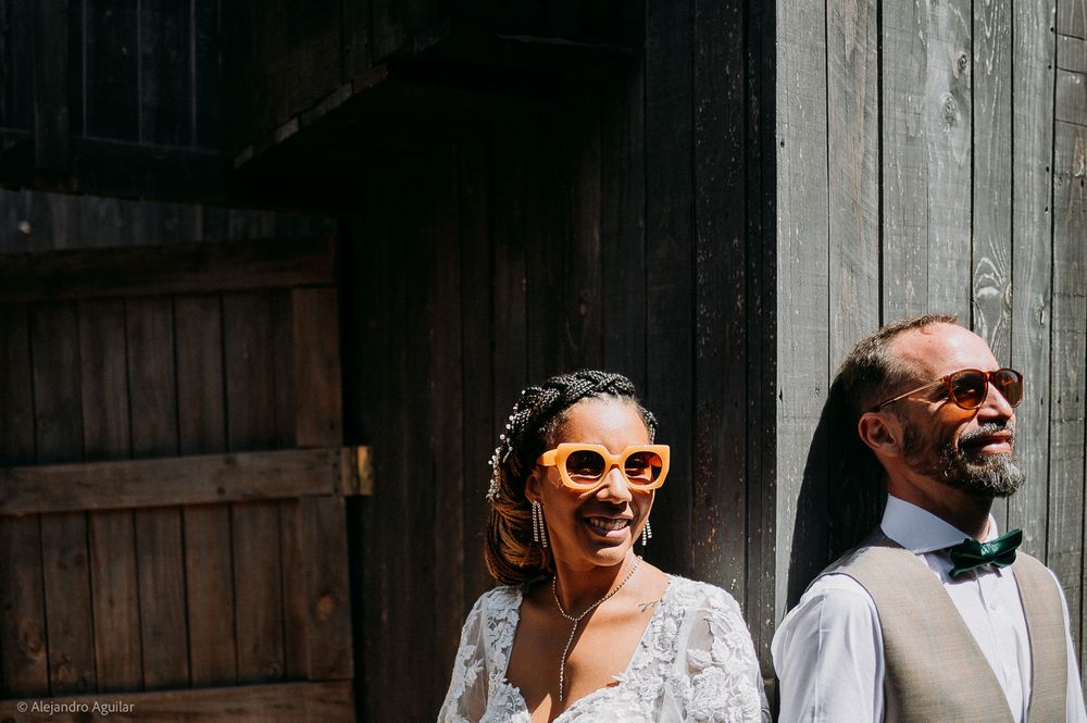 A smiling bride in a lace gown with yellow sunglasses stands beside a bearded groom in a vest near a rustic barn door.