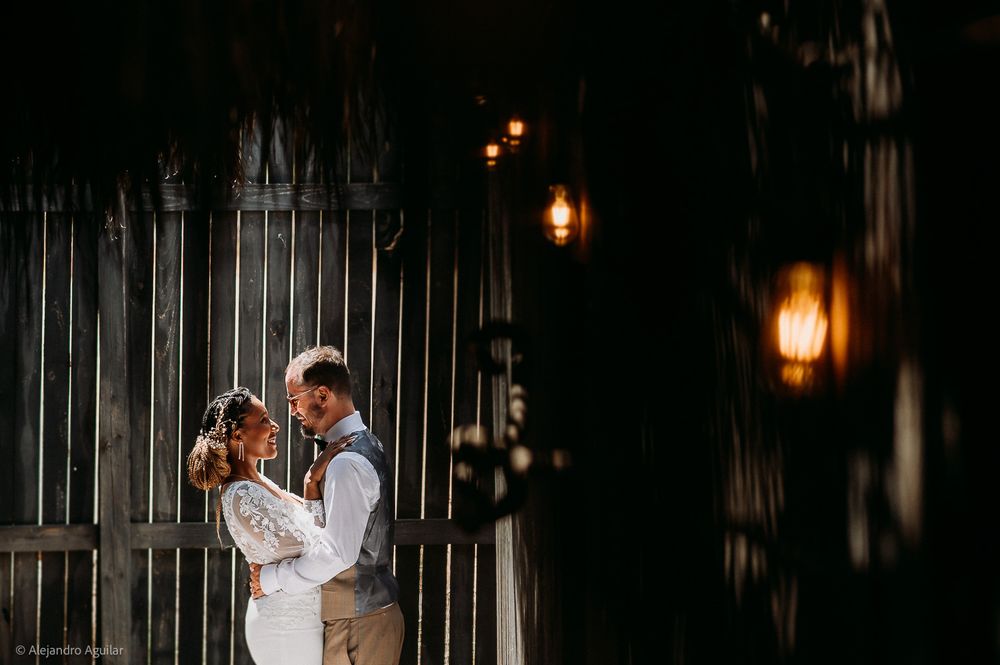 Romantic wedding couple embracing in front of rustic barn doors with warm glowing lanterns at night.