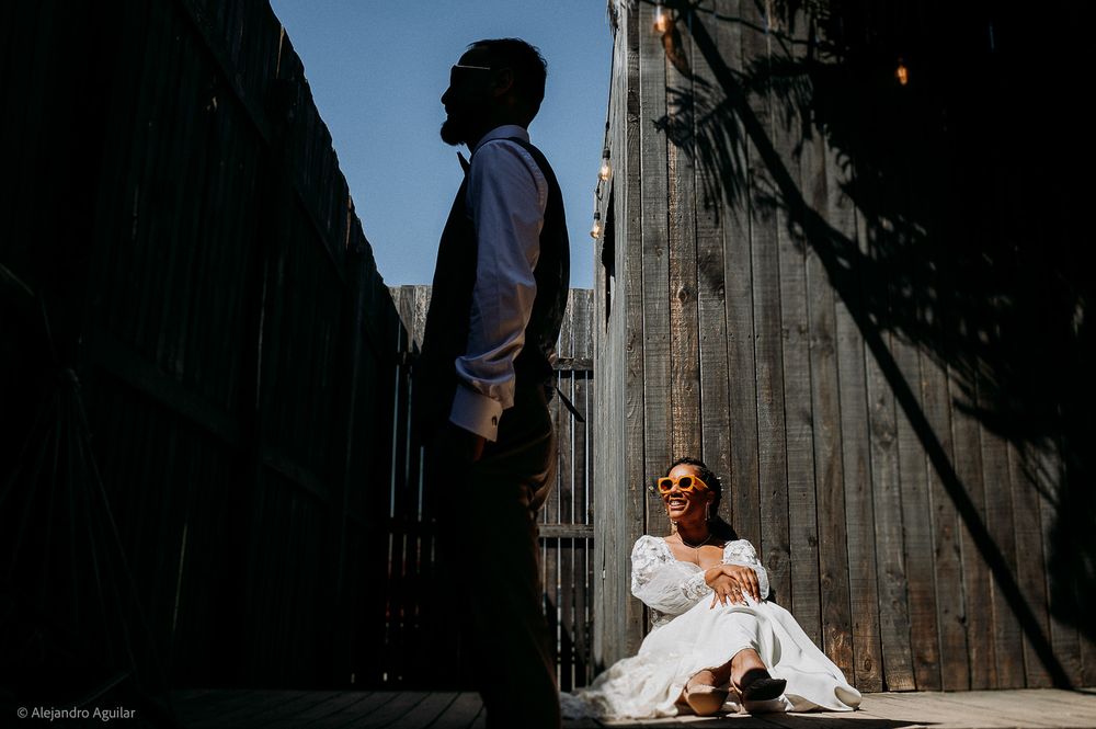 Couple in dramatic alley light, man standing in silhouette, woman in white dress seated against wooden fence.