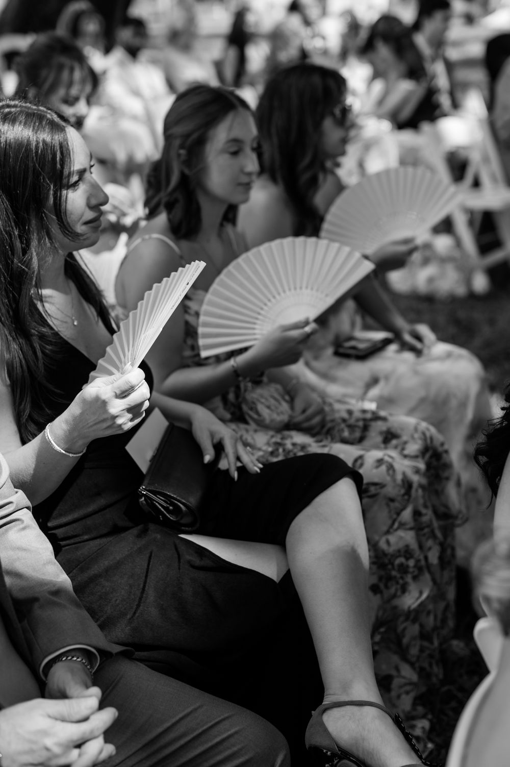 Black and white photo of wedding guests using white hand fans during an outdoor summer ceremony.