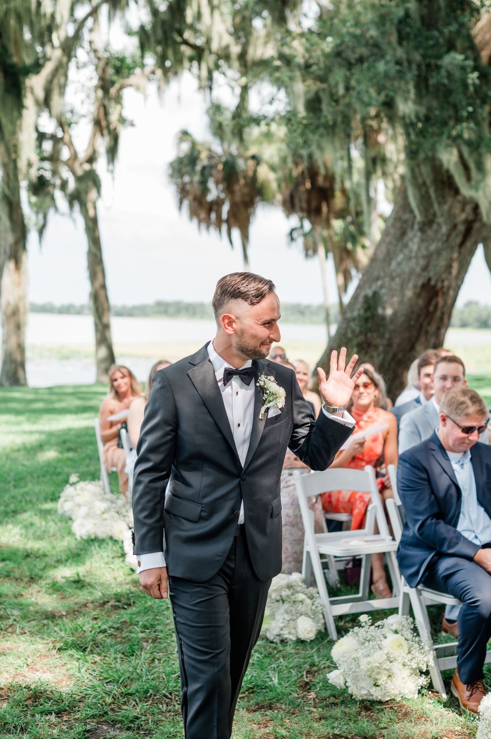 Dapper groom in black tuxedo waves to guests while walking down the outdoor aisle under moss-draped trees.