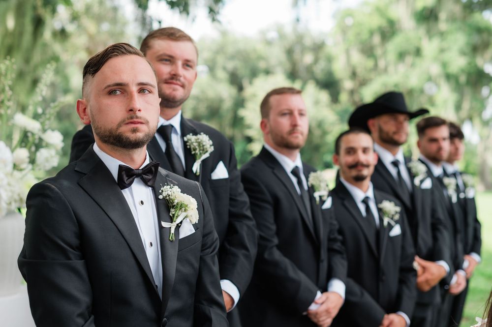 Groom and groomsmen in matching black tuxedos with boutonnieres stand attentively at outdoor wedding ceremony.