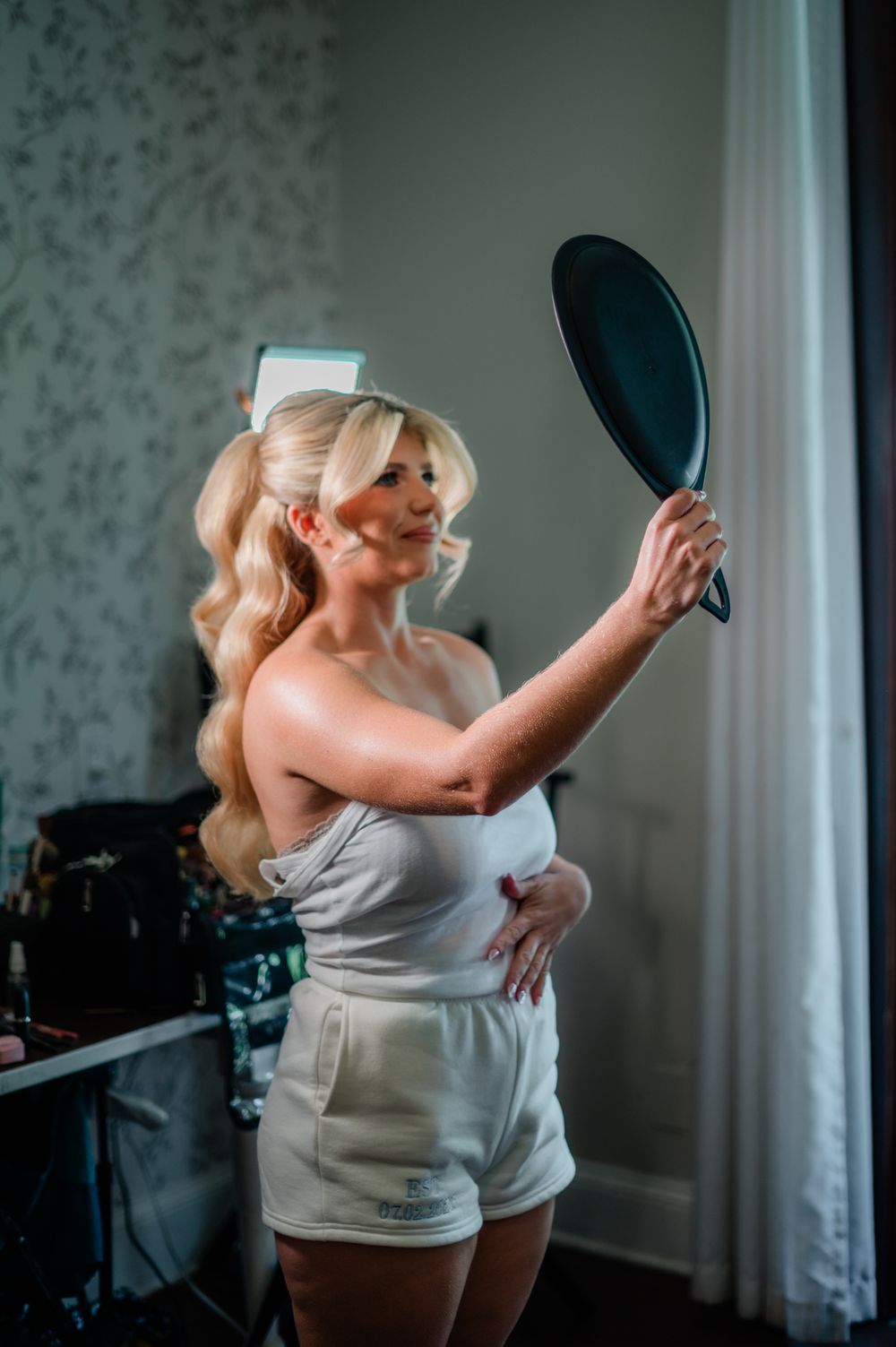Blonde woman in white outfit holding a hand mirror while getting ready in a softly lit room.