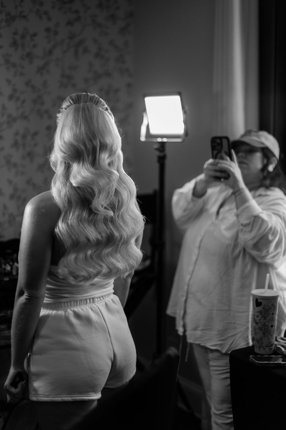Black and white photo of blonde woman with curly hair being photographed by person holding smartphone in mirror.
