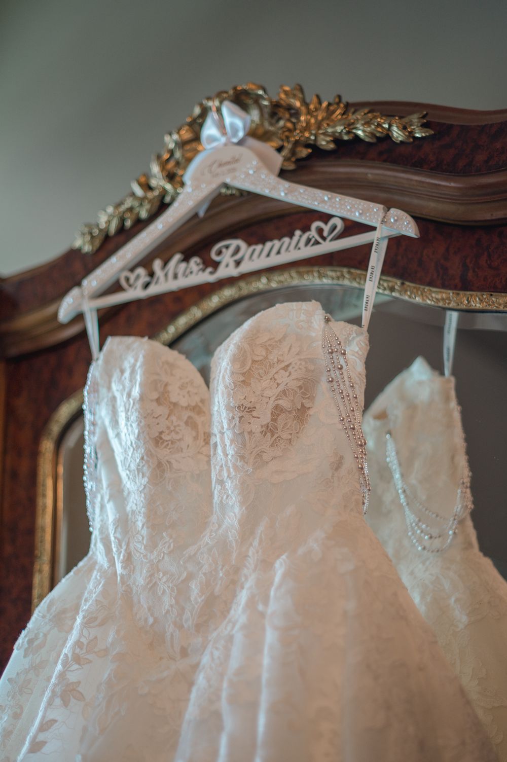White lace wedding dress hanging on a personalized Mrs. Ramie hanger on an ornate gold mirror frame.