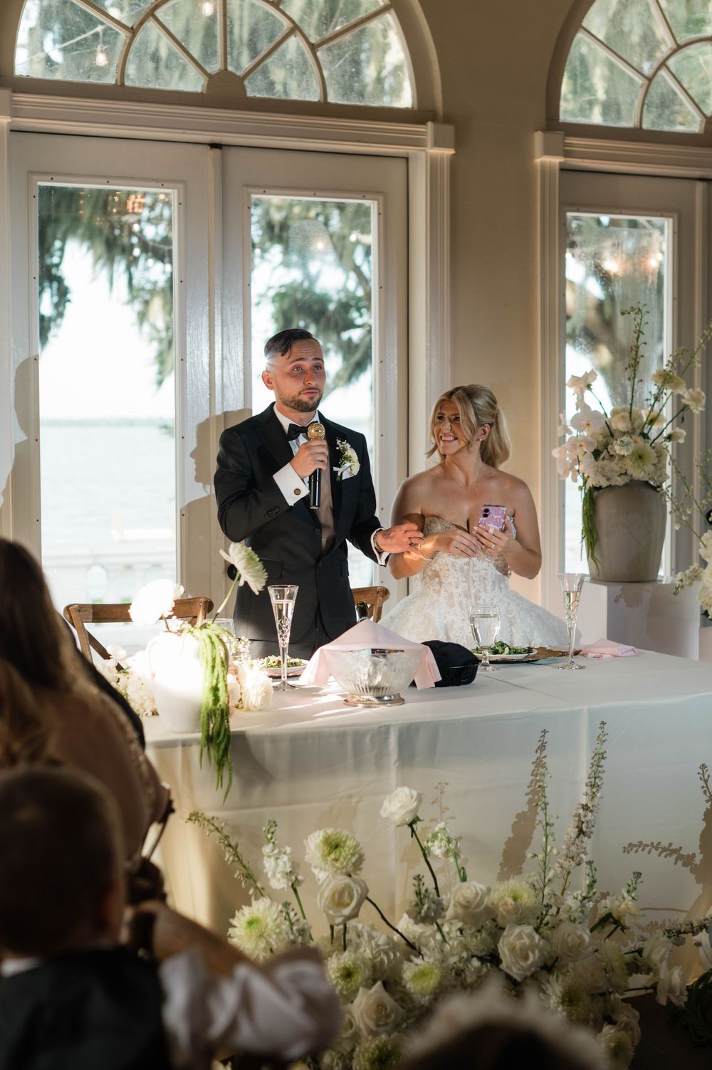 Bride and groom stand at sweetheart table giving speech at elegant wedding reception with white floral arrangements.