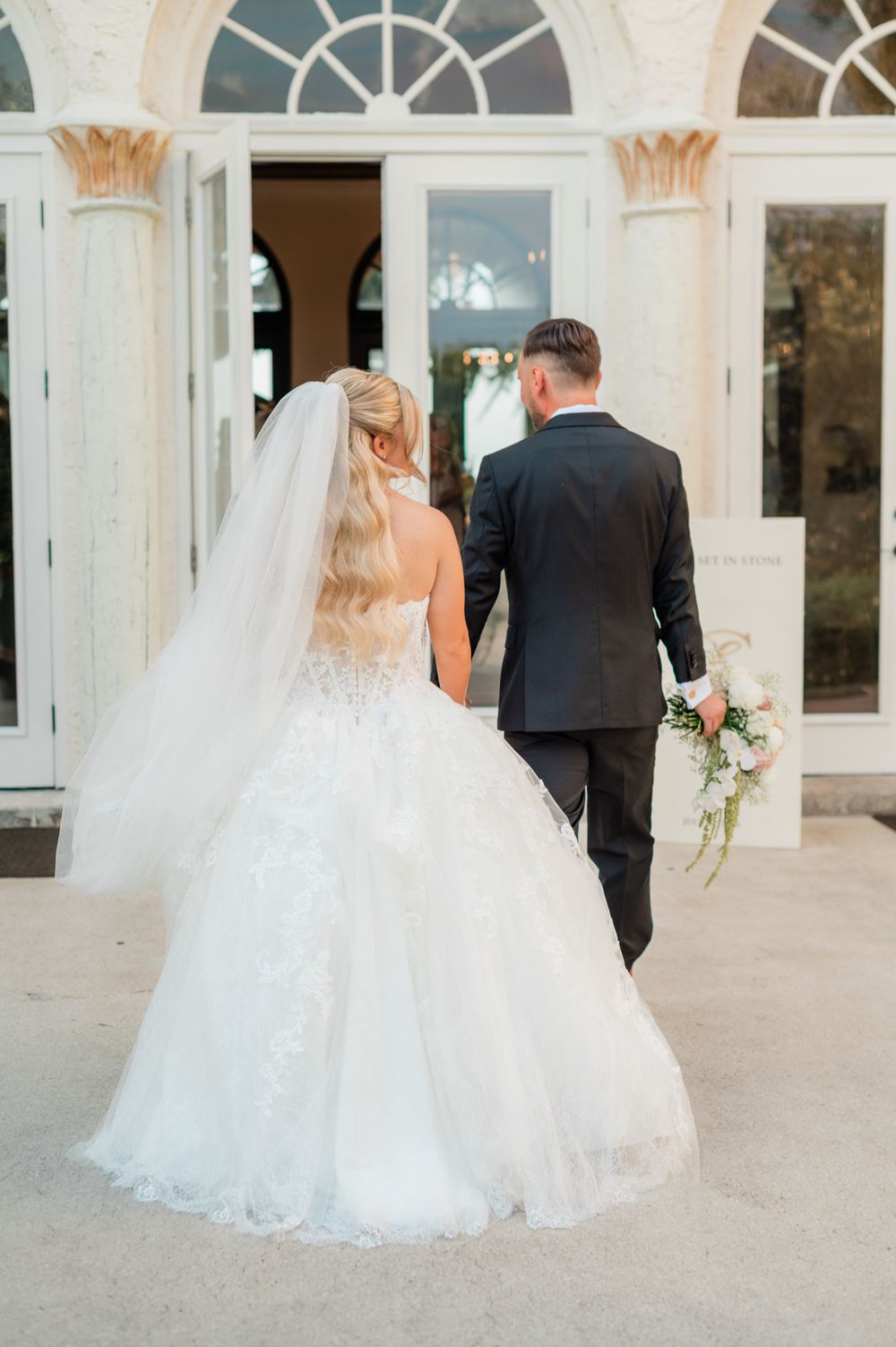 Bride in a lace ballgown with veil and groom in black tux walk hand-in-hand toward elegant white venue entrance.