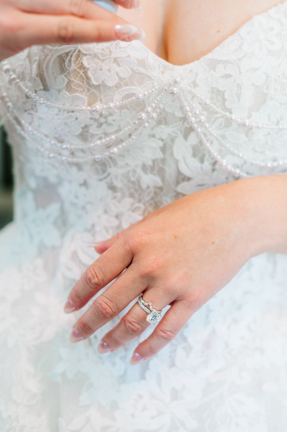 Close-up of bride's diamond engagement ring and wedding band on hand against intricate beaded lace wedding gown.
