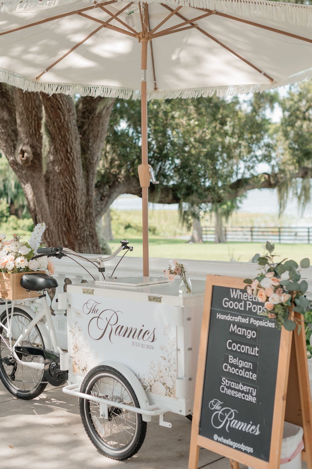Elegant white popsicle cart branded The Rumies with a chalkboard menu sign at an outdoor wedding reception under oak trees.