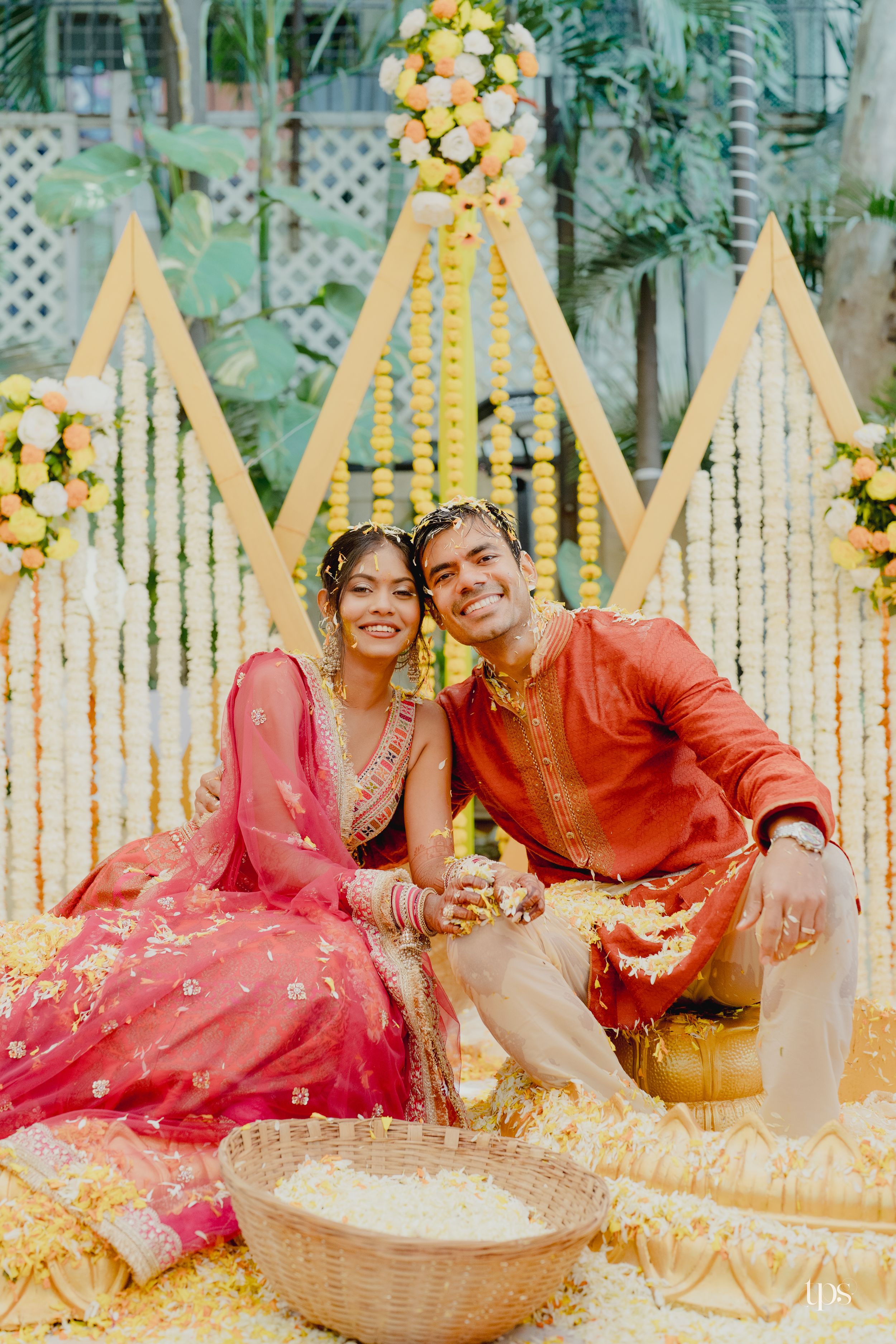 Indian couple in traditional red and orange attire sitting together during Haldi ceremony with floral backdrop.