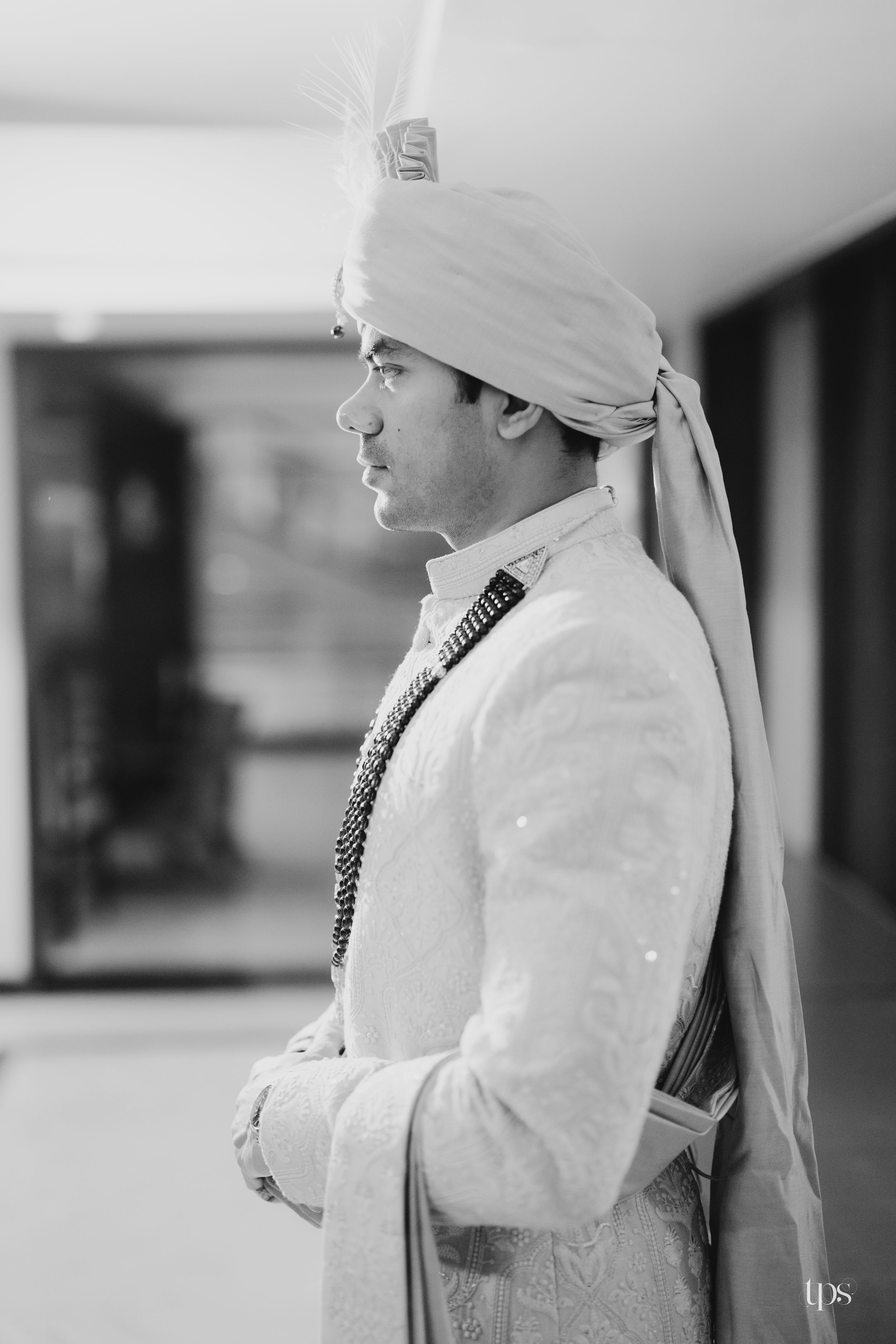 Black and white portrait of an Indian groom in traditional sherwani and turban with feather, standing with arms crossed.