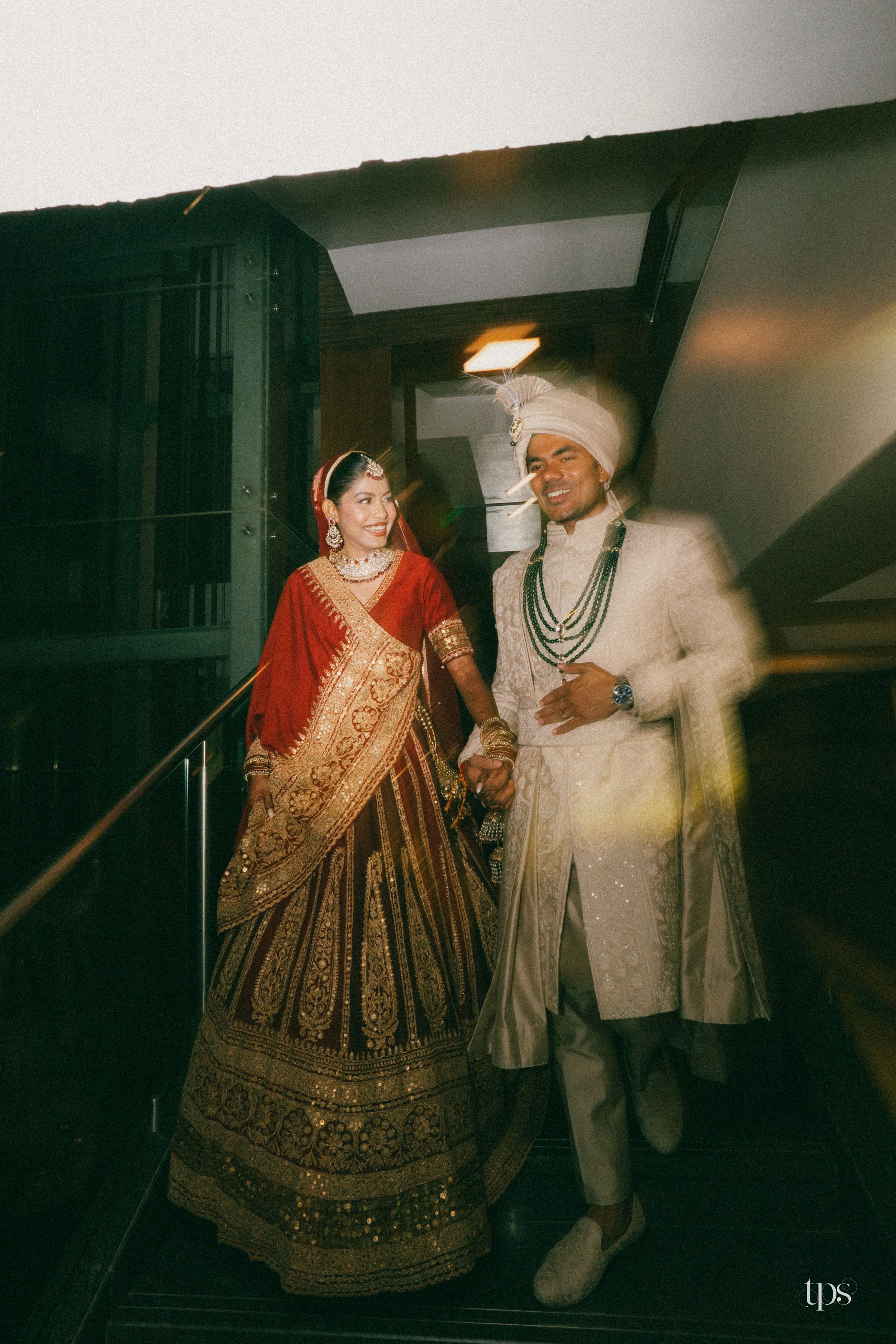 Indian bride in red lehenga and groom in white sherwani holding hands on stairs at traditional wedding ceremony.