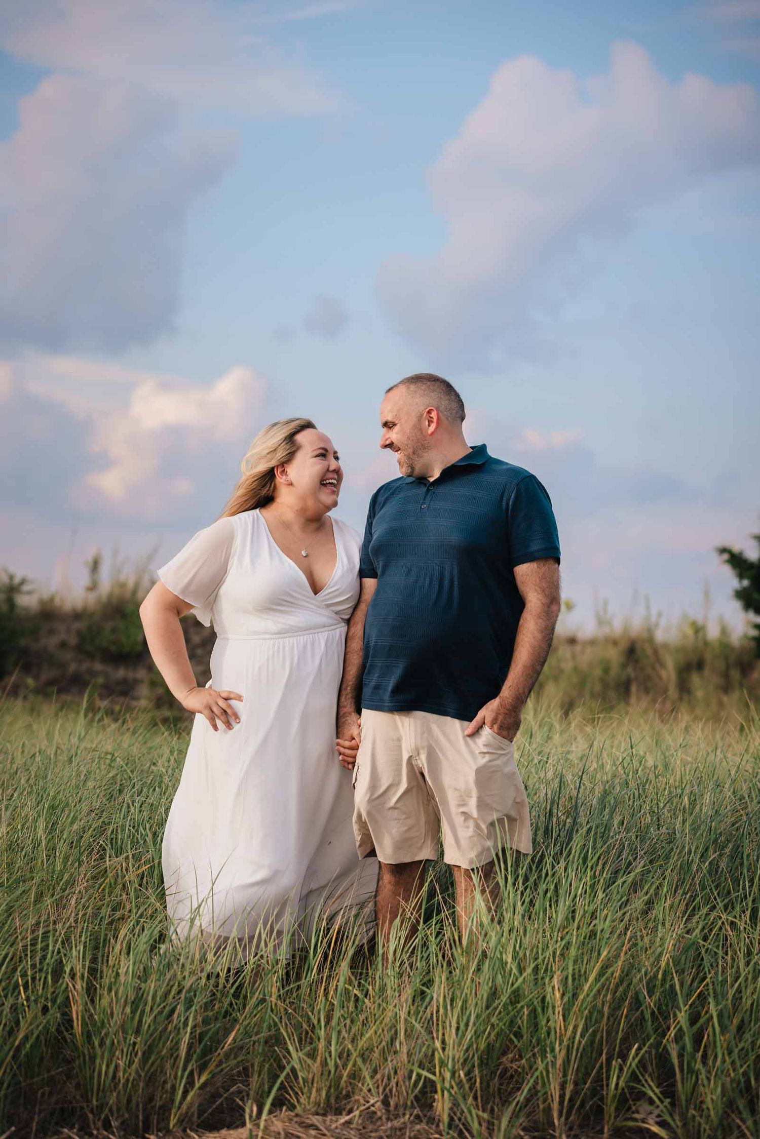 Engaged couple standing and laughing together in tall dune grass at Duxbury Beach, Massachusetts, beneath a blue sky.