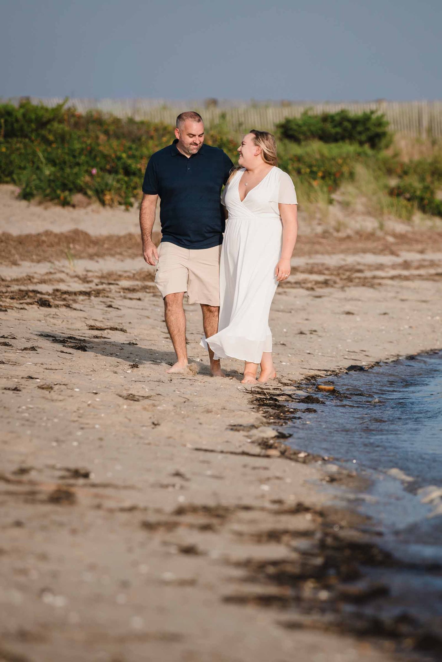 Engaged couple walking barefoot along the shoreline at Duxbury Beach, Massachusetts, smiling at each other in golden light.