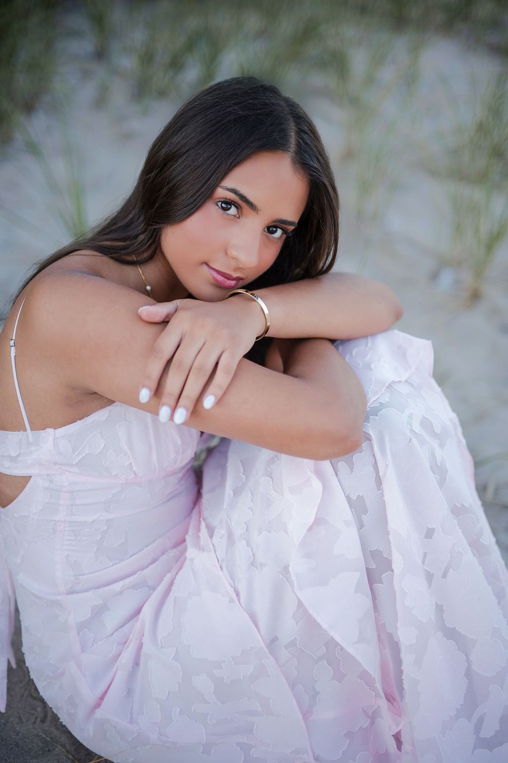 Young woman in pink floral dress sitting on beach sand, wearing gold bracelet and necklace, posing gracefully.