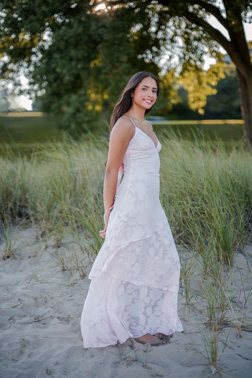 Young woman in a white lace maxi dress standing on sandy beach dunes with green grass and trees in background.
