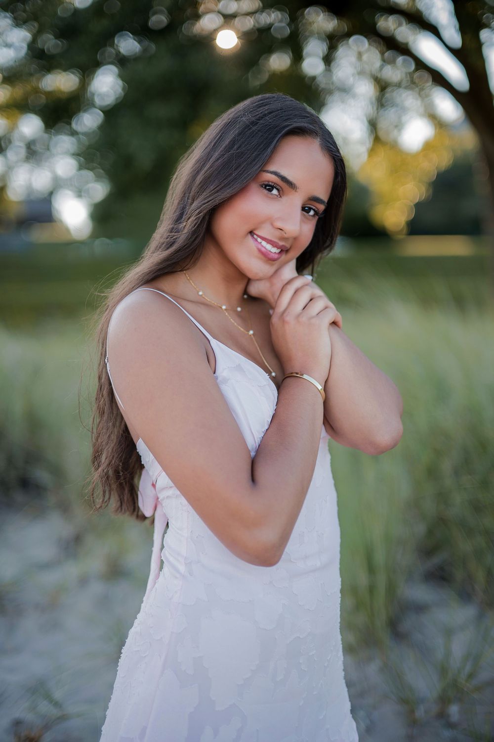 Young woman in white dress smiling outdoors, long brown hair, gold jewelry, natural green background.