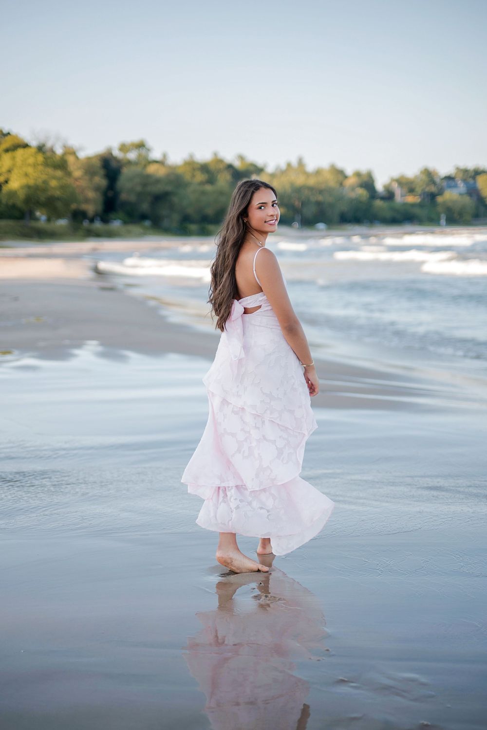 Young woman in a flowing pink dress walking barefoot on a sandy beach with ocean waves and trees in the background.