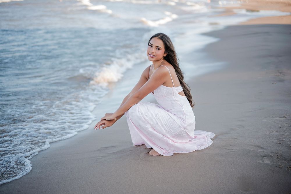Young woman in white dress sitting on sandy beach near ocean waves, smiling over shoulder at camera.