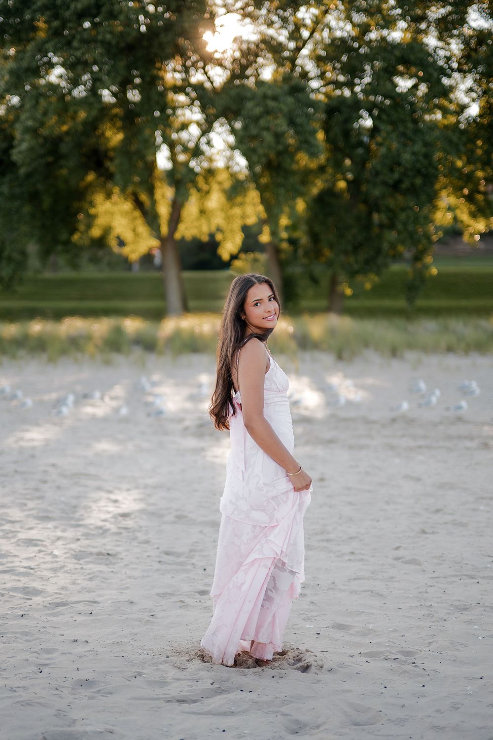 Young woman in pink dress standing on sandy beach at golden hour with green trees in background.