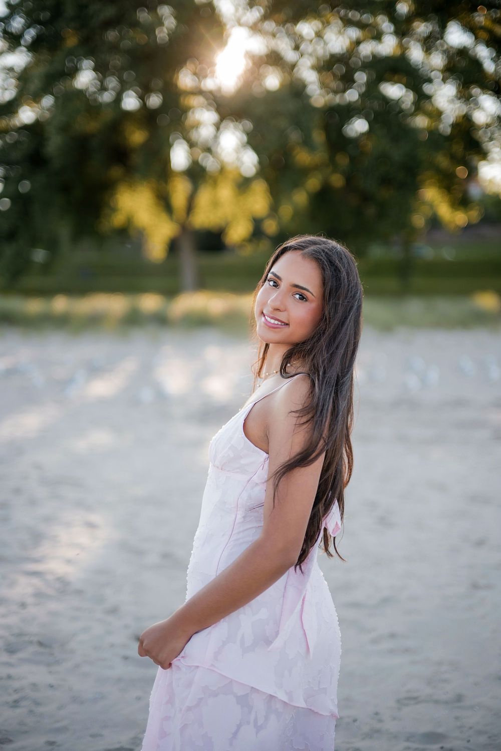 Young woman in white sundress smiling on sandy beach with sunlit trees in background.