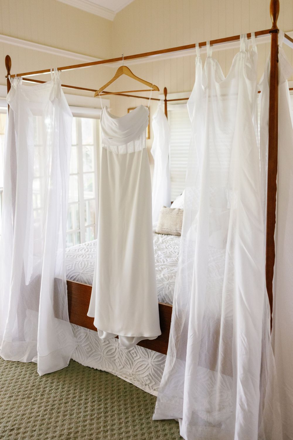 Elegant strapless white wedding gown hanging on a wooden hanger from a canopy bed frame.