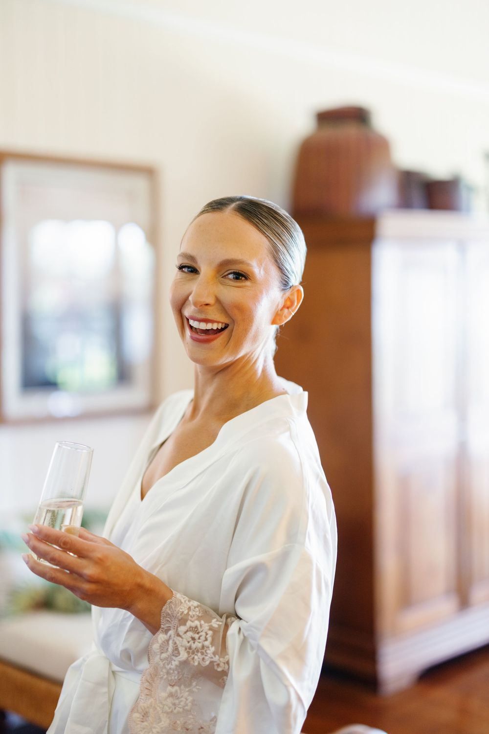 Radiant bride in white lace robe laughs joyfully while holding a champagne glass during wedding preparations.