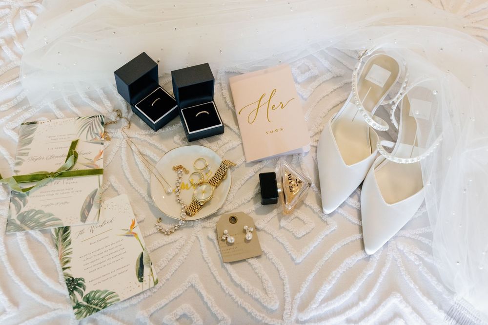 Bridal flat lay with white heels, gold jewelry, ring boxes, and wedding stationery on a white textured surface.