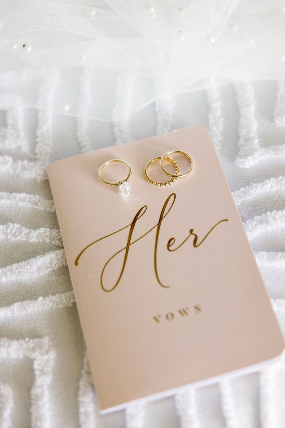 Gold wedding rings and diamond engagement ring resting on blush Her Vows booklet with elegant gold script.