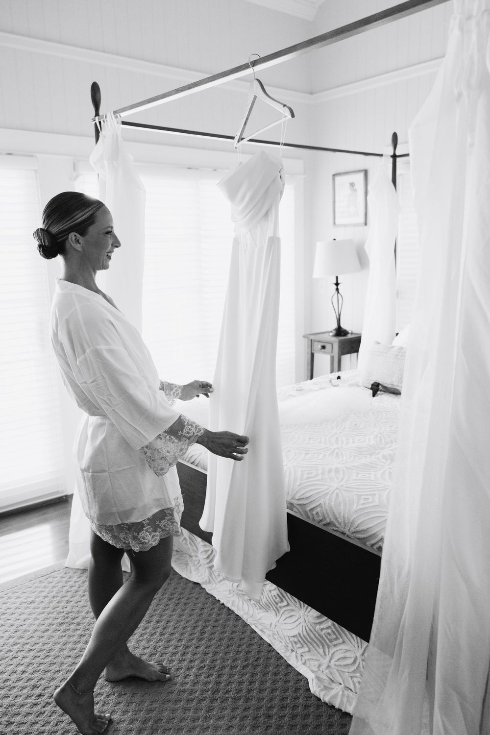 Bride in white robe admires her wedding gown hanging in a sunlit bedroom before the ceremony.