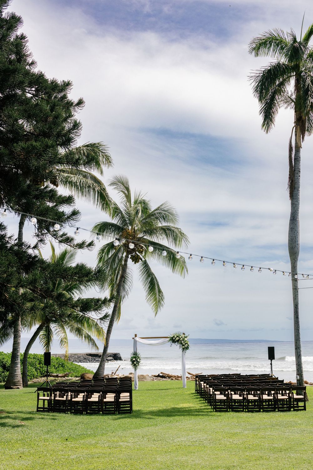 Outdoor beachside wedding ceremony setup with rows of chairs, floral arch, string lights, and palm trees overlooking the ocean.